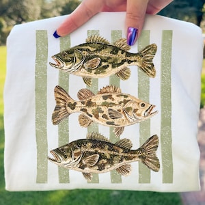 May include: White t-shirt featuring three detailed illustrations of fish in shades of green, brown, and tan. The fish are arranged vertically with a striped green background. The shirt is held up by a hand.