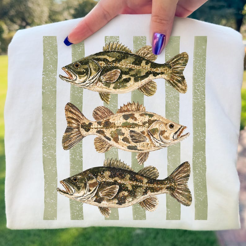 May include: White t-shirt featuring three detailed illustrations of fish in shades of green, brown, and tan. The fish are arranged vertically with a striped green background. The shirt is held up by a hand.