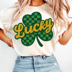 K&ouml;nnte beinhalten: Cremefarbenes T-Shirt mit einem gr&uuml;nen und gelben Kleeblatt-Design. Das Kleeblatt hat ein kariertes Muster und das Wort "Lucky" in gelber, kursiver Schrift. Das Shirt ist ein l&auml;ssiger Stil, passend zum St. Patrick's Day.