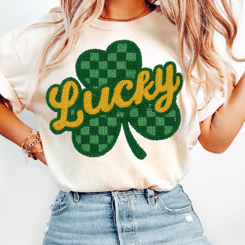 K&ouml;nnte beinhalten: Cremefarbenes T-Shirt mit einem gr&uuml;nen und gelben Kleeblatt-Design. Das Kleeblatt hat ein kariertes Muster und das Wort "Lucky" in gelber, kursiver Schrift. Das Shirt ist ein l&auml;ssiger Stil, passend zum St. Patrick's Day.