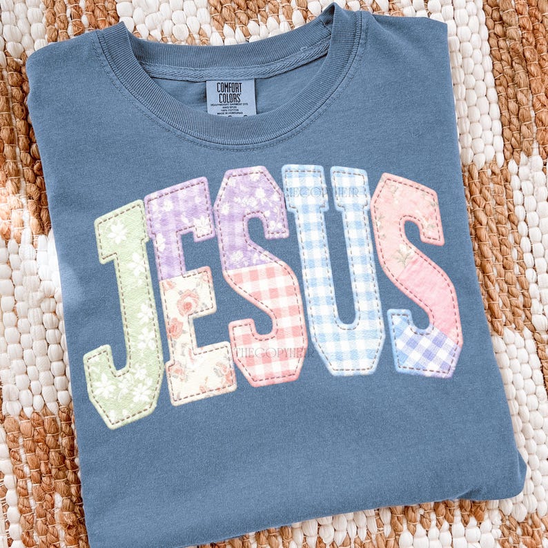 Puede incluir: Una camiseta azul Comfort Colors con la palabra "JESUS" aplicada en la parte delantera. Las letras son de estilo patchwork, utilizando telas florales, de cuadros y lisas en colores pastel. La camiseta est&aacute; doblada sobre una superficie tejida.