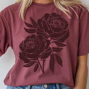 Puede incluir: Una camiseta de color granate con un diseño floral burdeos oscuro de dos peonías y hojas. El gráfico está centrado en la camiseta, creando una estética simple pero elegante. La camiseta tiene mangas cortas y cuello redondo.