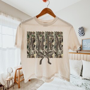 K&ouml;nnte beinhalten: Ein cremefarbenes T-Shirt mit dem Wort "AMEN" in einem Tarnmuster. Unter dem Wort befindet sich eine dunkelgraue Schleife. Das Shirt h&auml;ngt an einem Holzb&uuml;gel.