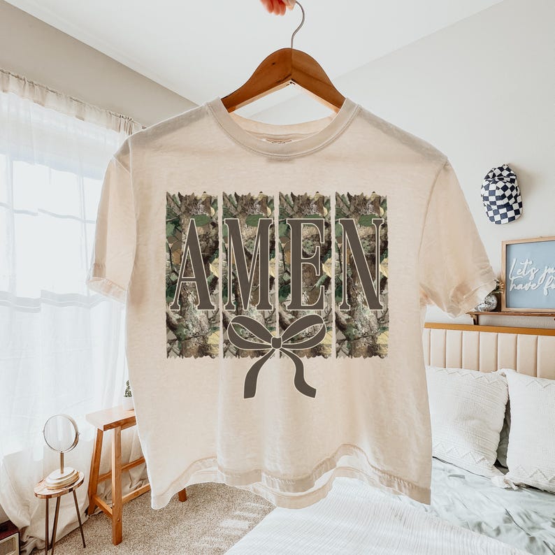 K&ouml;nnte beinhalten: Ein cremefarbenes T-Shirt mit dem Wort "AMEN" in einem Tarnmuster. Unter dem Wort befindet sich eine dunkelgraue Schleife. Das Shirt h&auml;ngt an einem Holzb&uuml;gel.