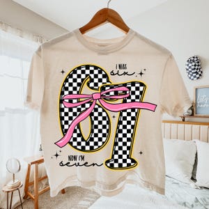 Op de afbeelding: Cr&egrave;mekleurig T-shirt met een groot "67"-motief in een zwart-wit geblokt patroon, geel omlijnd en versierd met een roze strik. De tekst "I WAS six" en "NOW I'M seven" zijn ook zichtbaar. Het shirt hangt aan een houten hanger.
