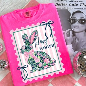 May include: A bright pink Comfort Colors sweatshirt featuring a floral bunny graphic with the words "Happy Easter." The bunny is decorated with pink and green flowers and a pink bow. The design is set within a pink and white striped frame.