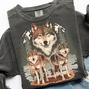 Puede incluir: Sudadera gris oscuro con una ilustraci&oacute;n detallada de tres lobos sobre un fondo de bosque y monta&ntilde;as. Los lobos est&aacute;n representados en tonos marrones, grises y blancos, con una est&eacute;tica vintage. La sudadera tiene cuello redondo y mangas largas.