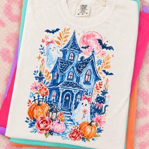 May include: A cream-colored t-shirt featuring a graphic print of a blue haunted house, two ghosts with pink bows, a black cat, orange pumpkins, flowers, bats, and a pink moon. The shirt has a Comfort Colors brand label at the inner neckline.