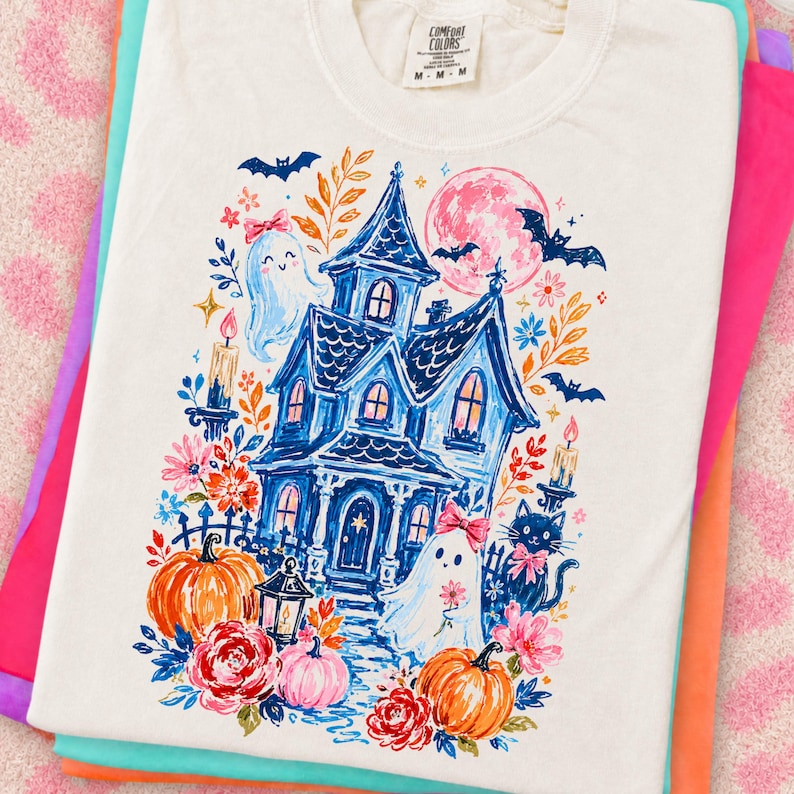 May include: A cream-colored t-shirt featuring a graphic print of a blue haunted house, two ghosts with pink bows, a black cat, orange pumpkins, flowers, bats, and a pink moon. The shirt has a Comfort Colors brand label at the inner neckline.
