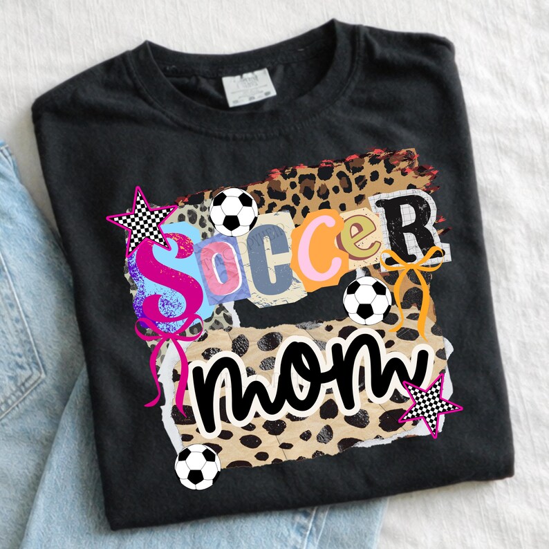 May include: Black t-shirt with a colorful graphic that says "Soccer Mom." The design includes soccer balls, stars, and a leopard print background. The word "Soccer" is in a collage style with different colors.