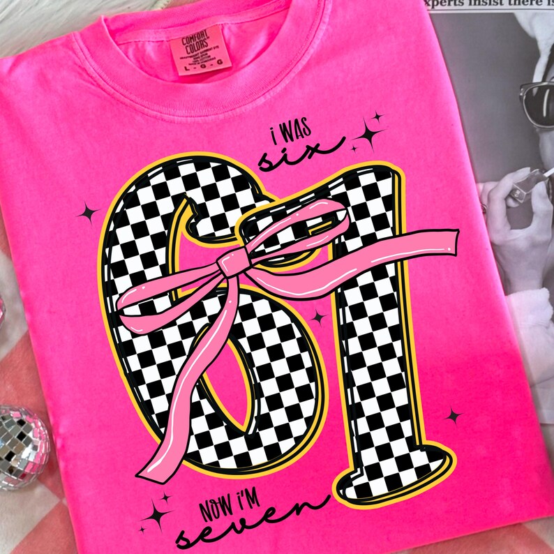 Op de afbeelding: Een felroze t-shirt met de tekst "I WAS six, NOW I'M seven." Het getal "61" staat centraal, ontworpen met een zwart-wit geblokt patroon en een roze strik. Het shirt heeft een ronde hals.