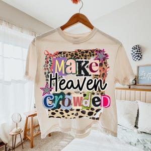 May include: A cream-colored crop top with the phrase "Make Heaven Crowded" in colorful, block letters. The text is set against a leopard print background with star accents. The shirt is hanging on a wooden hanger.