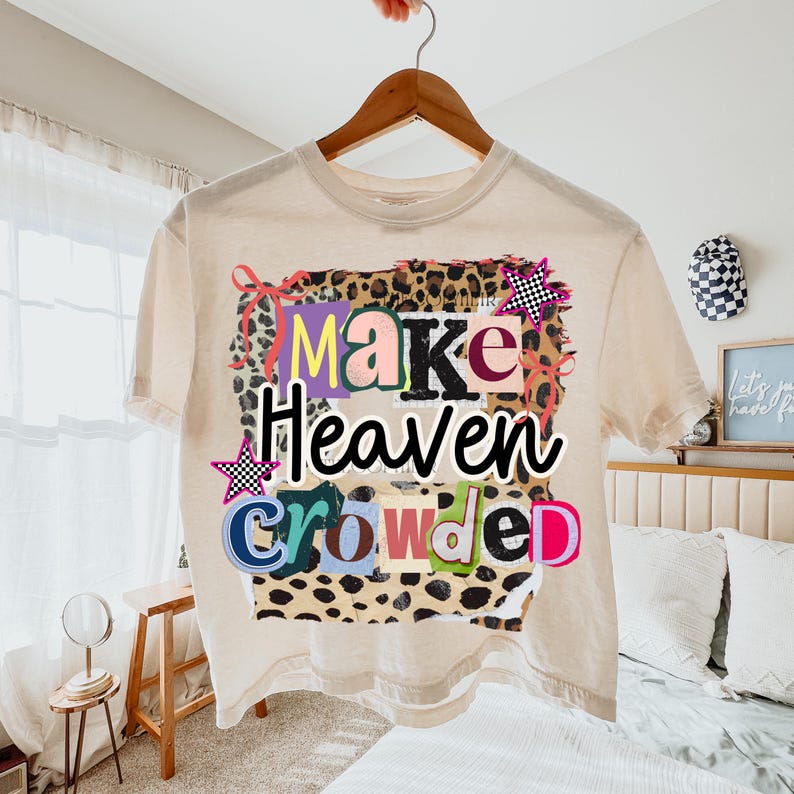 May include: A cream-colored crop top with the phrase "Make Heaven Crowded" in colorful, block letters. The text is set against a leopard print background with star accents. The shirt is hanging on a wooden hanger.