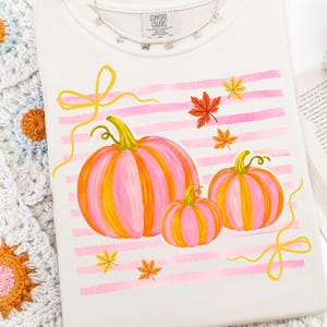 May include: Cream-colored t-shirt featuring a watercolor design of three pumpkins in shades of pink and orange, with yellow stems and leaves. Pink horizontal stripes and yellow ribbons accent the design. A delicate silver necklace with small charms is at the top.