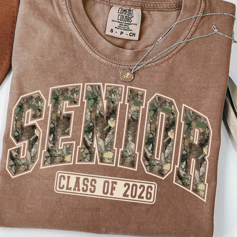 Pu&ograve; includere: Una maglietta Comfort Colors marrone con la scritta "SENIOR" a grandi lettere con motivo mimetico. Sotto, il testo "CLASS OF 2026" &egrave; stampato in un riquadro bianco. Una collana d'argento con un ciondolo &egrave; appoggiata sulla maglietta.