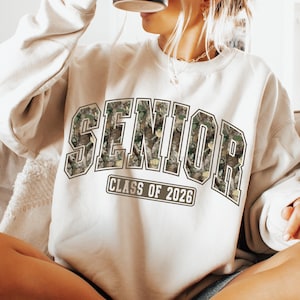 Pu&ograve; includere: Felpa color crema con la scritta "SENIOR" a grandi lettere con motivo mimetico. Sotto c'&egrave; la scritta "CLASS OF 2026". Questa felpa casual &egrave; adatta per la laurea ed &egrave; un capo comodo.