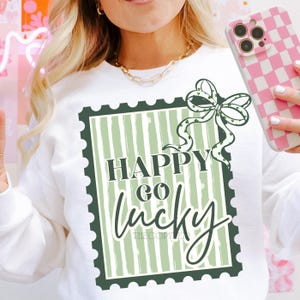 Puede incluir: Sudadera blanca con un gr&aacute;fico verde y blanco que dice "Happy Go Lucky" en un dise&ntilde;o de sello. Un lazo verde con tr&eacute;boles est&aacute; encima del texto. La sudadera es de estilo casual.