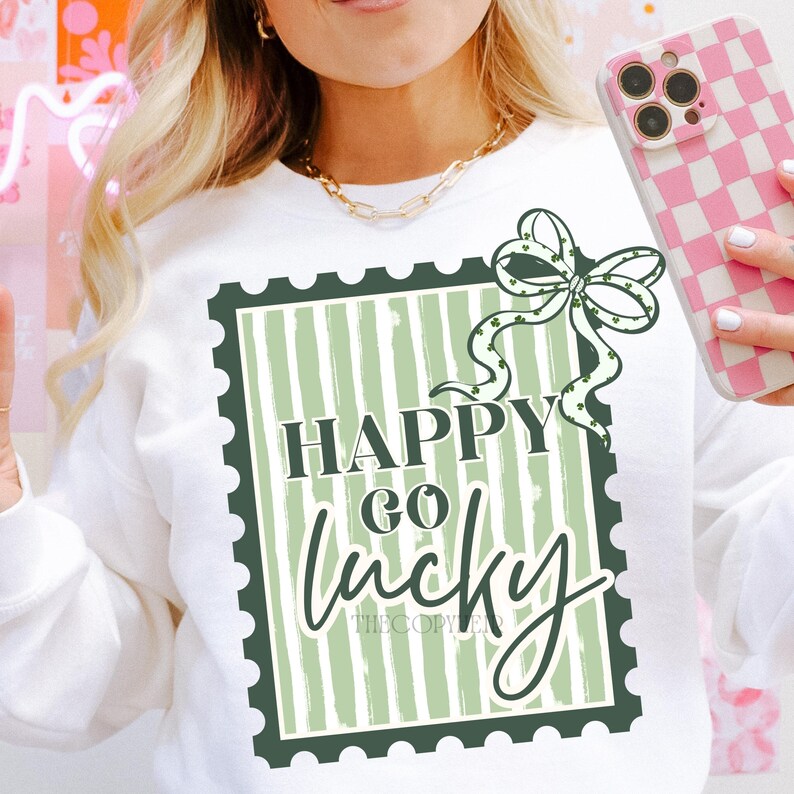 Puede incluir: Sudadera blanca con un gr&aacute;fico verde y blanco que dice "Happy Go Lucky" en un dise&ntilde;o de sello. Un lazo verde con tr&eacute;boles est&aacute; encima del texto. La sudadera es de estilo casual.