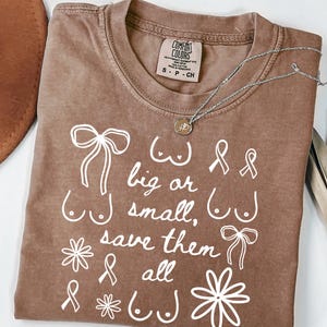 May include: A brown Comfort Colors t-shirt with white text and illustrations. The text reads "big or small, save them all" with illustrations of breasts, bows, and ribbons. A silver necklace with a pendant is on the shirt.