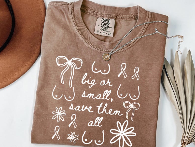 May include: A brown Comfort Colors t-shirt with white text and illustrations. The text reads "big or small, save them all" with illustrations of breasts, bows, and ribbons. A silver necklace with a pendant is on the shirt.