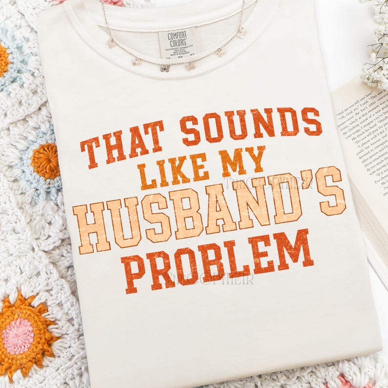May include: Cream-colored t-shirt with the phrase "THAT SOUNDS LIKE MY HUSBAND'S PROBLEM" in orange and peach lettering. The shirt is folded and displayed with a delicate butterfly necklace and a crocheted blanket.
