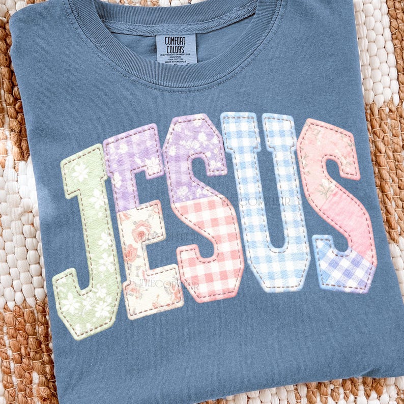 May include: A blue Comfort Colors t-shirt with the word "JESUS" in a patchwork design. The letters are in various pastel floral and gingham patterns, including green, purple, pink, and blue. The shirt is folded on a woven surface.