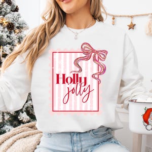 Puede incluir: Sudadera blanca con un dise&ntilde;o festivo. El dise&ntilde;o presenta las palabras "Holly Jolly" en escritura roja dentro de un marco a rayas rosas y blancas, coronado por un lazo rosa. Un art&iacute;culo de ropa con tem&aacute;tica navide&ntilde;a.
