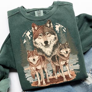 Puede incluir: Una sudadera verde oscuro con un gr&aacute;fico de tres lobos en tonos marr&oacute;n y gris, sobre un fondo de bosque y monta&ntilde;as. La sudadera tiene cuello redondo y mangas largas. El nombre de la marca "COMFORT COLORS" es visible.