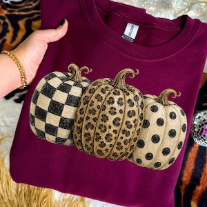 May include: A maroon sweatshirt featuring three decorative pumpkins. The pumpkins have unique patterns: a black and white checkerboard, a leopard print, and black polka dots. The Gildan brand tag is visible.