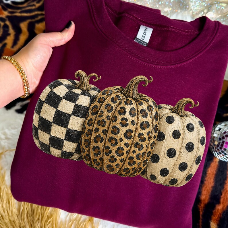 May include: A maroon sweatshirt featuring three decorative pumpkins. The pumpkins have unique patterns: a black and white checkerboard, a leopard print, and black polka dots. The Gildan brand tag is visible.