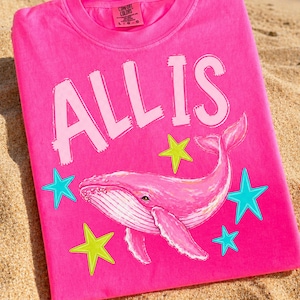 May include: A bright pink t-shirt with the words "ALL IS" in white block letters. A pink whale illustration is centered below the text, surrounded by teal and yellow star graphics. The shirt is made of a soft material.