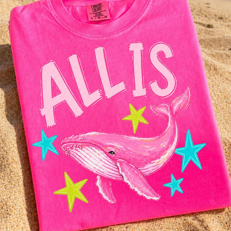 May include: A bright pink t-shirt with the words "ALL IS" in white block letters. A pink whale illustration is centered below the text, surrounded by teal and yellow star graphics. The shirt is made of a soft material.