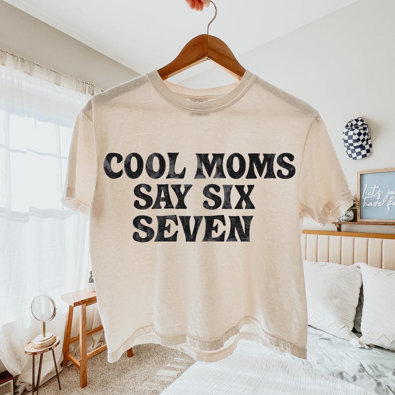 May include: Cream-colored t-shirt with the text "COOL MOMS SAY SIX SEVEN" in bold, black letters. The shirt is hanging on a wooden hanger. The style is casual and relaxed, suitable for everyday wear.