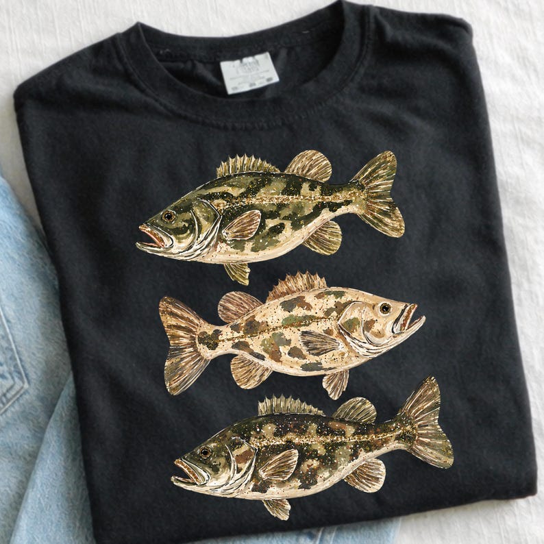 May include: Black t-shirt featuring three detailed illustrations of fish in shades of green, brown, and tan. The fish are arranged vertically, showcasing their scales and fins. The shirt is made of a soft material.