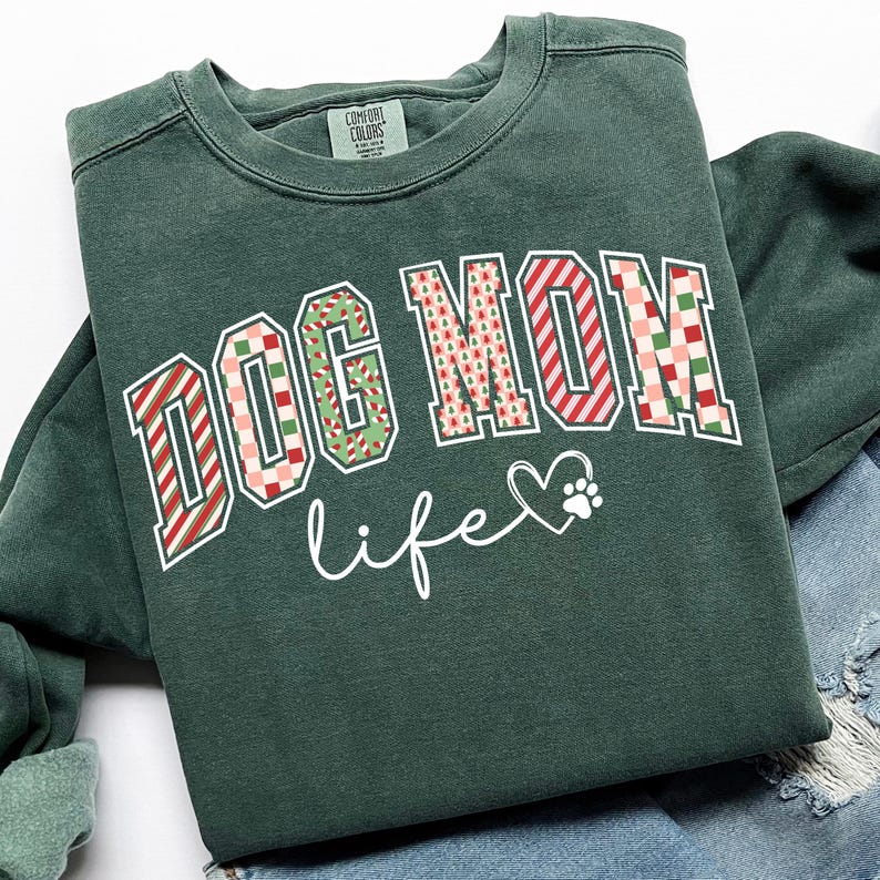 Może przedstawiać: Ciemnozielona bluza z napisem "DOG MOM life" w ozdobnej czcionce. Litery "DOG MOM" mają świąteczny wz&oacute;r w kolorze czerwonym, zielonym i białym. Obok słowa "life" znajduje się serce i odcisk łapy.