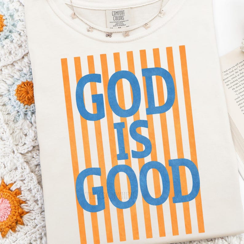 Puede incluir: Camiseta color crema con el texto "GOD IS GOOD" en letras azules, sobre un fondo de rayas verticales naranjas. Un delicado collar de mariposas descansa sobre la camiseta. La camiseta es de Comfort Colors.