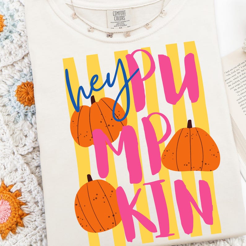 May include: Cream-colored t-shirt with the words "hey PUMPKIN" in blue and pink, with orange pumpkin graphics. The shirt has yellow vertical stripes in the background. A delicate necklace with small charms is visible at the top.