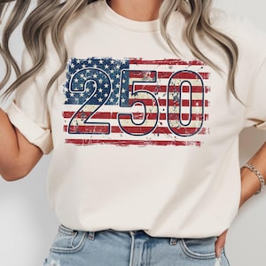 Puede incluir: Camiseta color crema con un dise&ntilde;o desgastado de la bandera estadounidense y el n&uacute;mero "250" en azul y rojo. El gr&aacute;fico est&aacute; centrado en la camiseta, con un aspecto vintage y desgastado. La camiseta est&aacute; hecha de un material suave.