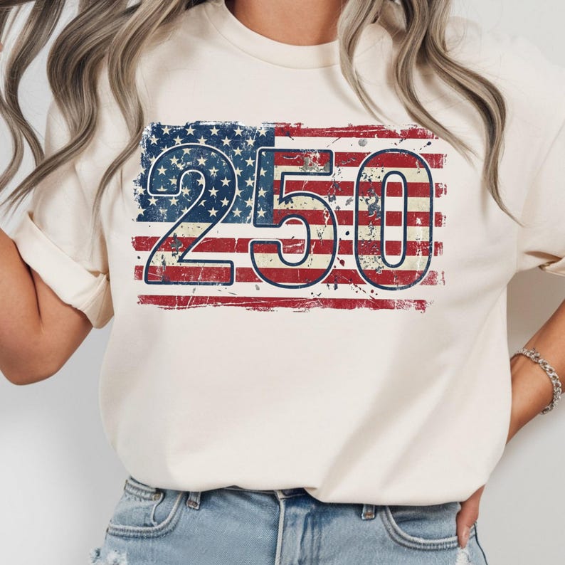 Puede incluir: Camiseta color crema con un dise&ntilde;o desgastado de la bandera estadounidense y el n&uacute;mero "250" en azul y rojo. El gr&aacute;fico est&aacute; centrado en la camiseta, con un aspecto vintage y desgastado. La camiseta est&aacute; hecha de un material suave.