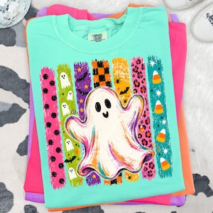 May include: A mint green crew neck t-shirt featuring a large central graphic of a white ghost. The design includes vertical brushstroke stripes in various colours and patterns, including black polka dots, small ghosts, bats, orange checkers, spiderwebs, and candy corn.