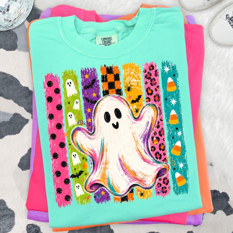 May include: A mint green crew neck t-shirt featuring a large central graphic of a white ghost. The design includes vertical brushstroke stripes in various colours and patterns, including black polka dots, small ghosts, bats, orange checkers, spiderwebs, and candy corn.