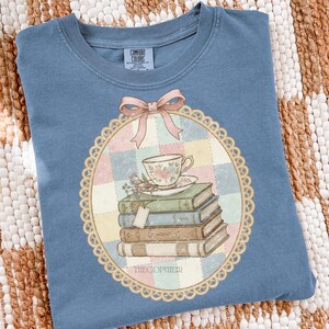 May include: A blue Comfort Colors t-shirt with a detailed graphic. The graphic features a teacup, books, and a bow. The design is framed by a scalloped border and the text "THECOPYHEIR" is at the bottom.