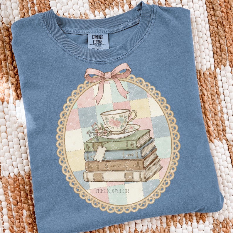 May include: A blue Comfort Colors t-shirt with a detailed graphic. The graphic features a teacup, books, and a bow. The design is framed by a scalloped border and the text "THECOPYHEIR" is at the bottom.