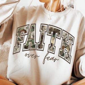 May include: Cream-colored sweatshirt with the word "FAITH" in large, block letters filled with a camouflage pattern. Below the word "FAITH" is the phrase "over fear" in a cursive font. The sweatshirt is a casual style.