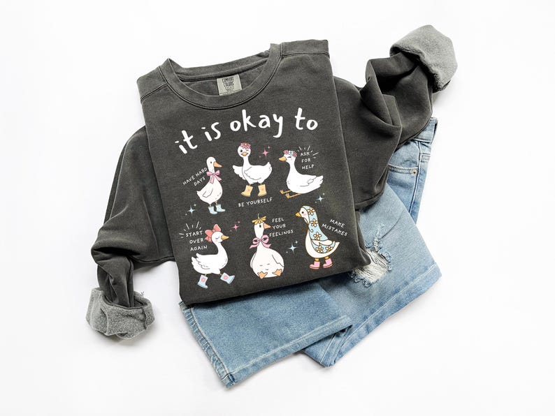 Pode incluir: Camisola cinza escuro com a frase "it is okay to" e ilustra&ccedil;&otilde;es de gansos. Os gansos usam acess&oacute;rios e t&ecirc;m bal&otilde;es de texto com afirma&ccedil;&otilde;es positivas. A camisola &eacute; combinada com cal&ccedil;&otilde;es de ganga azul claro.