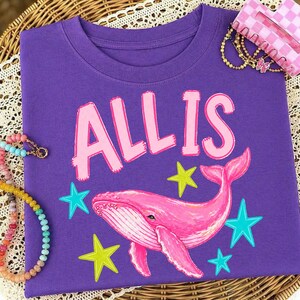 May include: A purple t-shirt featuring the name "ALLIS" in pink letters above a pink whale illustration. The design includes blue and yellow star accents. The shirt is displayed on a woven surface with colorful beaded necklaces.