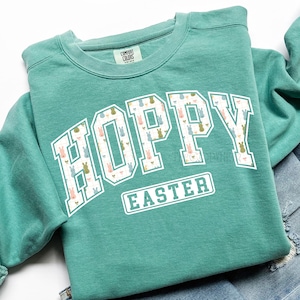 May include: A teal green sweatshirt with the word "HOPPY" in large white letters filled with a bunny pattern. Below the word "HOPPY" is the word "EASTER" in a white rectangular box. The sweatshirt has a crew neck and long sleeves.