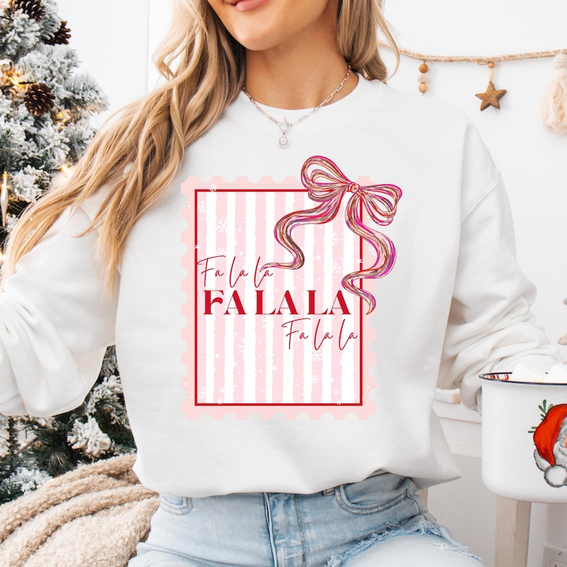 Puede incluir: Sudadera blanca con un dise&ntilde;o de rayas rosas y blancas. El dise&ntilde;o incluye un lazo rosa y el texto "Fa la la FALALA Fa la la" en un marco rojo. Una prenda festiva.