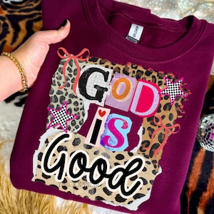 Może przedstawiać: Bordowa bluza z okrągłym dekoltem z kolorową grafiką z napisem "GOD IS GOOD". Projekt zawiera nadruk w cętki, gwiazdy i wstążki. Swobodny, wygodny styl ubioru.