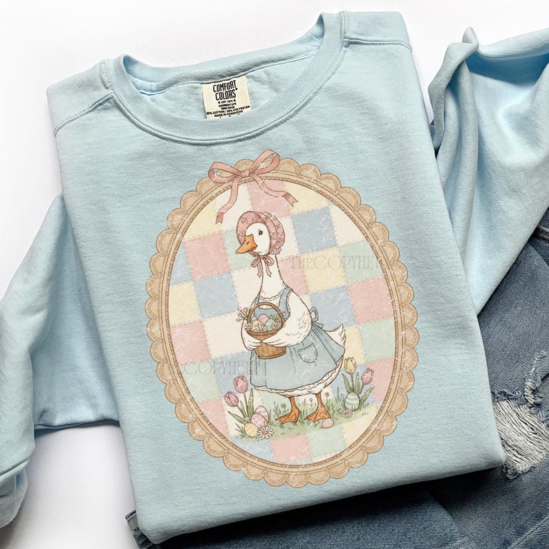 Puede incluir: Sudadera Comfort Colors azul claro con una ilustraci&oacute;n ovalada de un ganso con gorro y delantal, llevando una cesta de huevos de Pascua. La ilustraci&oacute;n est&aacute; sobre un fondo de patchwork pastel con tulipanes y el texto "THECOPYHE".